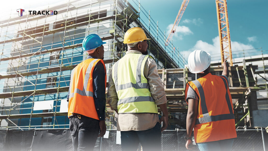 Three construction workers in orange vests and hard hats stand at a scaffolded building, discussing progress near a construction site logo in the corner.