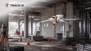 Drone with orange lights hovering over a messy construction site, surveying equipment nearby and a Track3D logo in the corner.