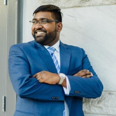 Smiling man in a blue suit with a striped tie, arms crossed, standing against a light wall outside.