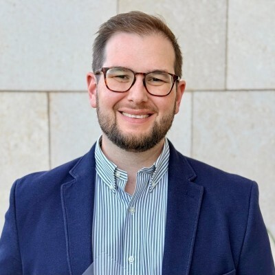 Head-and-shoulders portrait of a smiling man with glasses, wearing a navy blazer and striped shirt, standing against a light stone wall.