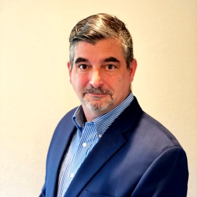 Professional head-and-shoulders portrait of a man in a navy blazer and blue striped shirt, standing against a light wall with a neutral expression.