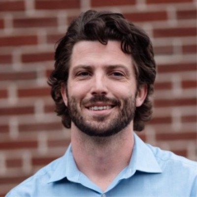 Smiling man with dark hair and beard in a light blue shirt, standing in front of a brick wall.