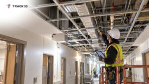 Site lead inspecting pipes and ductwork above an open ceiling grid in a hospital corridor under construction